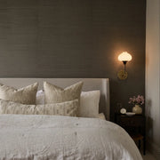 Modern wall light fixture mounted above a bedside table with a glowing glass shade and brass backplate.