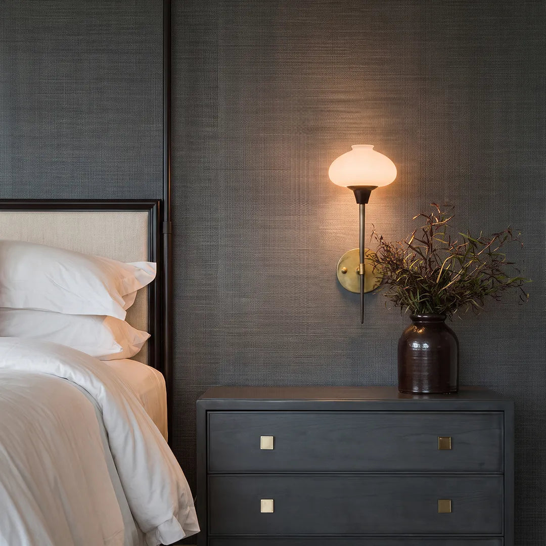 Modern wall light fixture beside a bed with a dark textured wall and warm ambient glow.