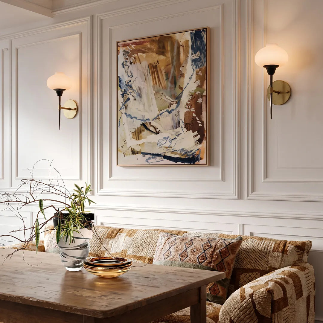 Modern wall light fixture installed on a white paneled wall beside abstract artwork and a sofa.
