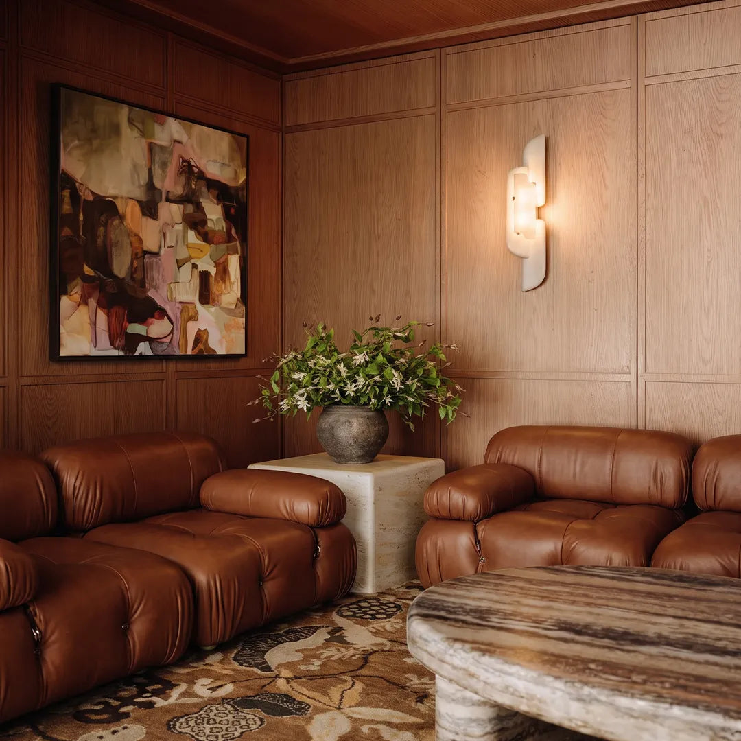 Modern wall light fixtures illuminating a living room with brown leather sofas and wood-paneled walls.