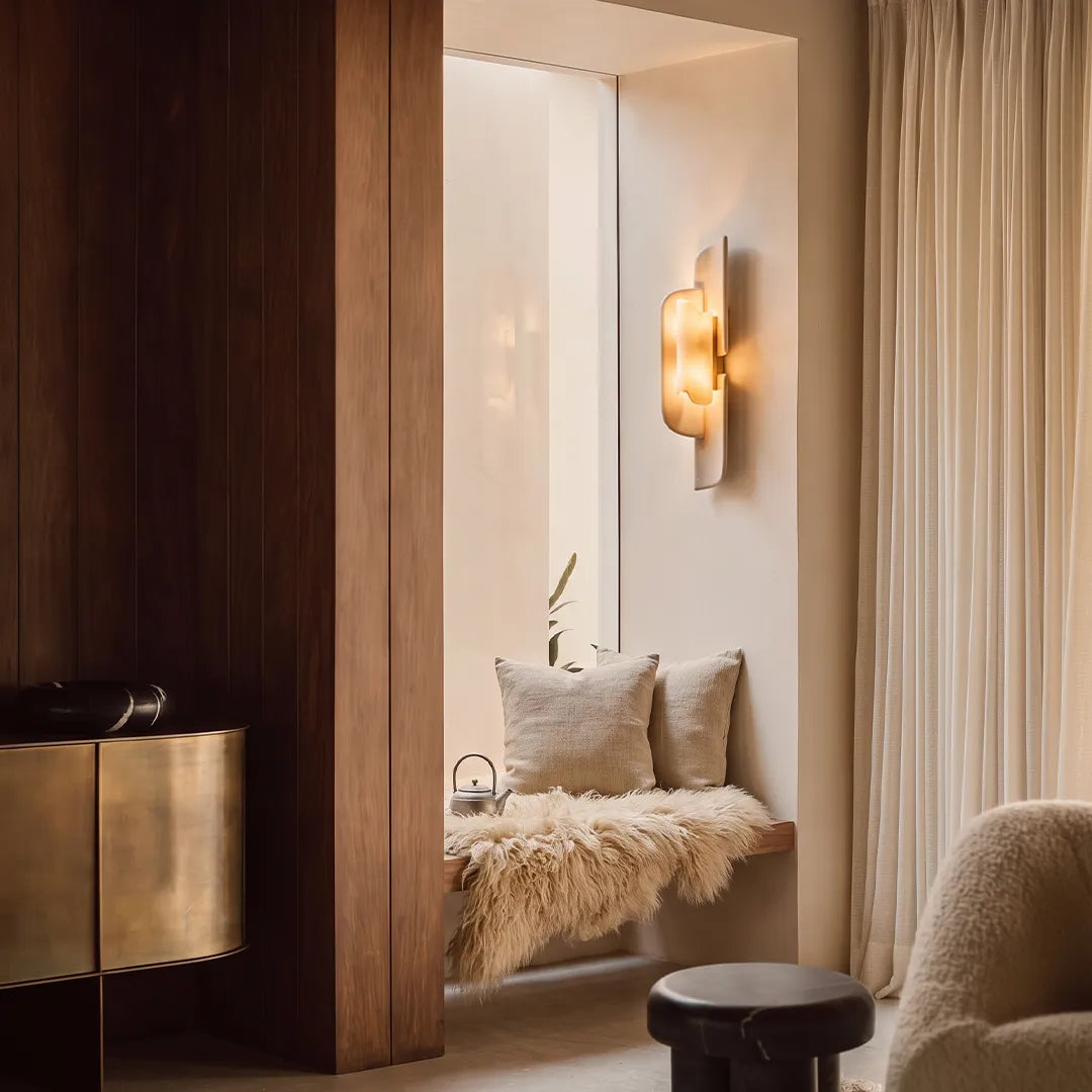 Modern wall light fixtures mounted beside a cozy window seat in a warm neutral interior.