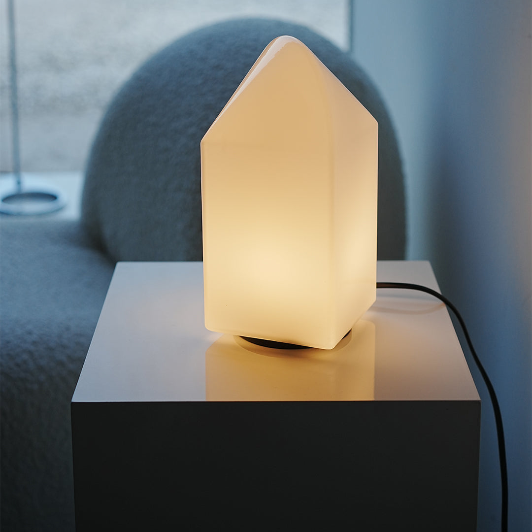 Small modern white table lamp house-shaped placed on bedside table, cozy ambient glow.