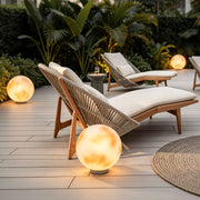 Moon-shaped stone light illuminating a poolside deck near wicker lounge seating in a tropical outdoor setting.