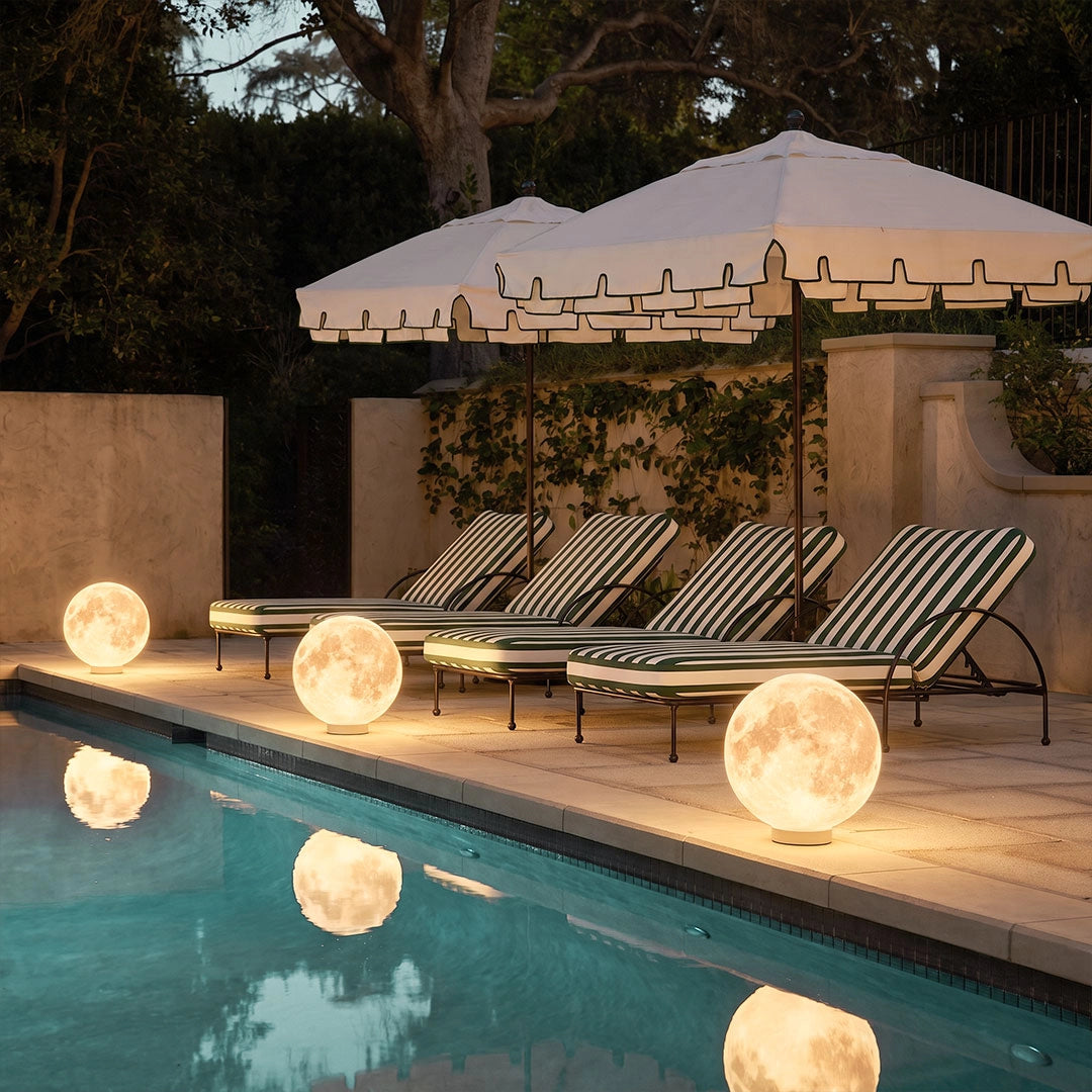 Moon-shaped stone light glowing beside a swimming pool and lounge chairs in an outdoor patio setting at night.