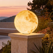 Moon-shaped stone pillar light illuminating garden pathway with warm glow at sunset.