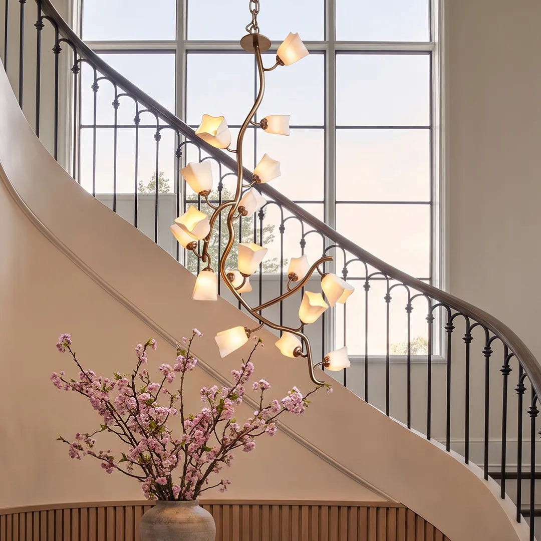 Multi light pendant chandelier suspended beside a curved staircase in a bright foyer with tall windows.