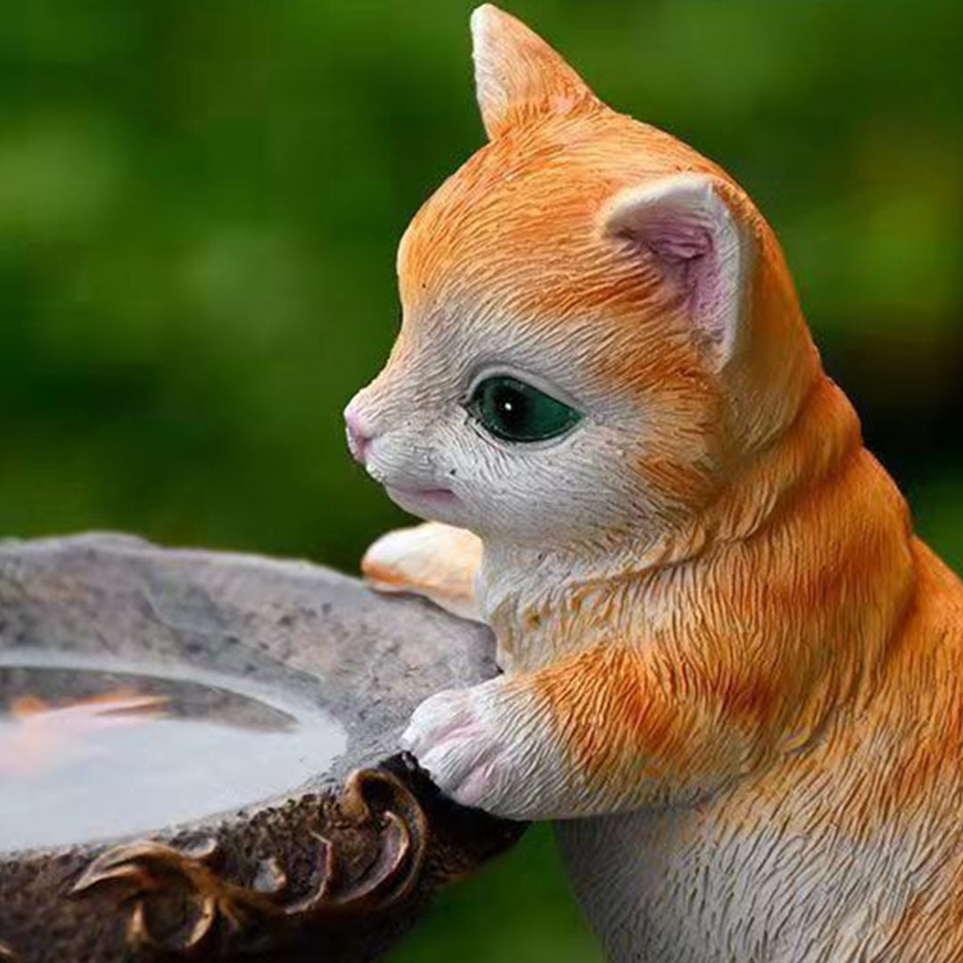 Orange tabby kitten figurine positioned on ornate fountain rim in natural garden environment