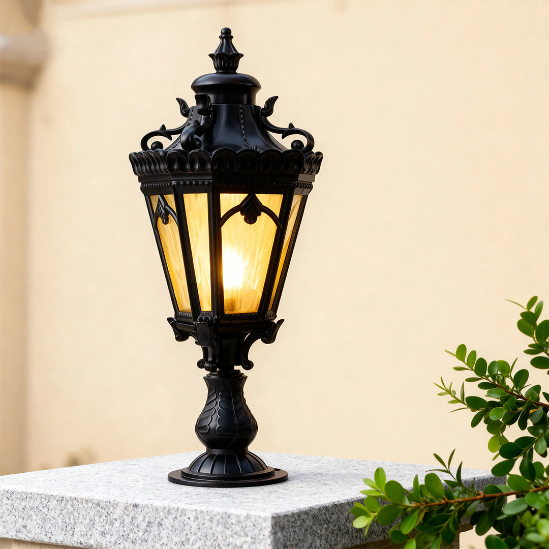 Ornate black outdoor pillar light brightening beige column with pink and coral flowering trees behind