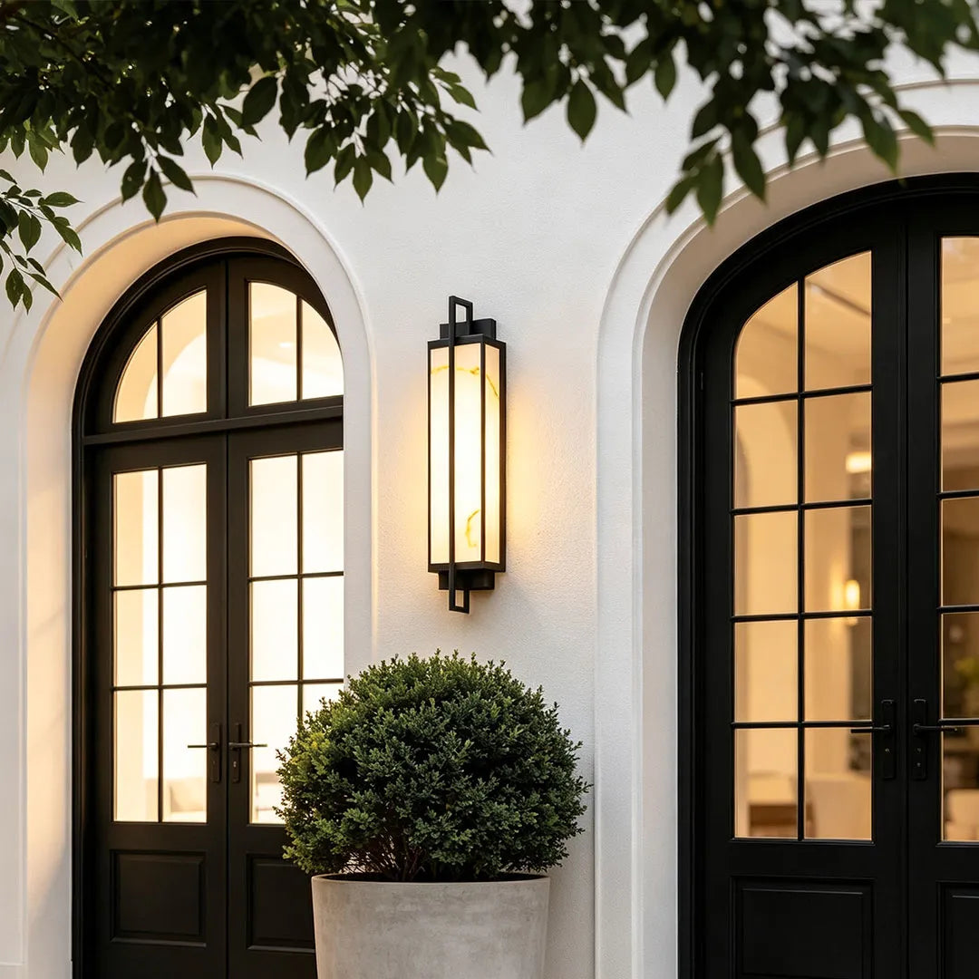 Outdoor alabaster wall sconce mounted between arched black framed doors, adding warm vertical accent lighting to a white exterior wall.