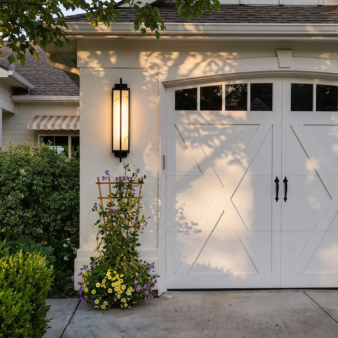 Outdoor alabaster wall sconce mounted beside a white garage door with flowers and soft evening light.