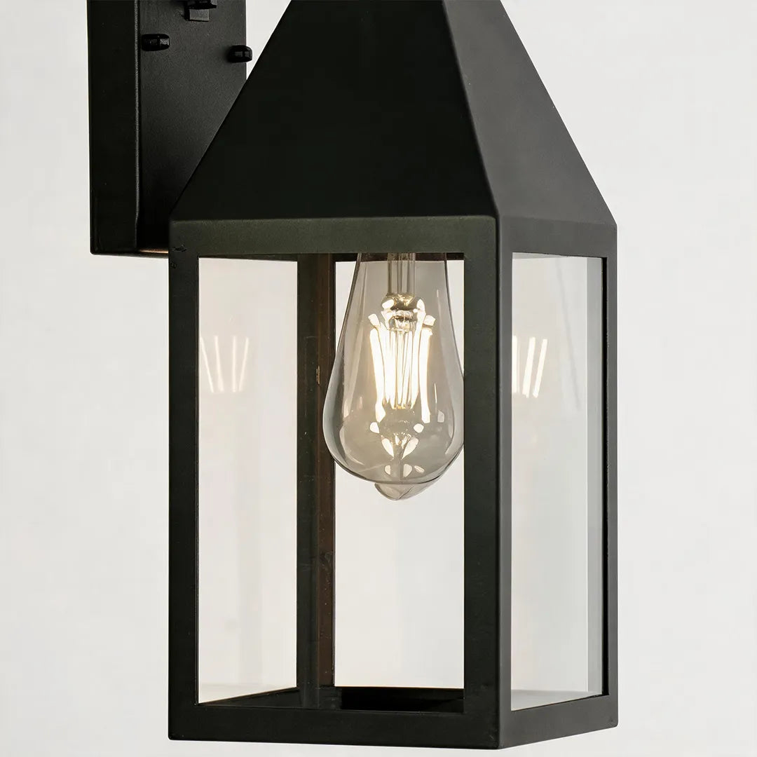 Outdoor black sconce close-up showing the tapered lantern frame, clear glass panels, and exposed bulb detail.