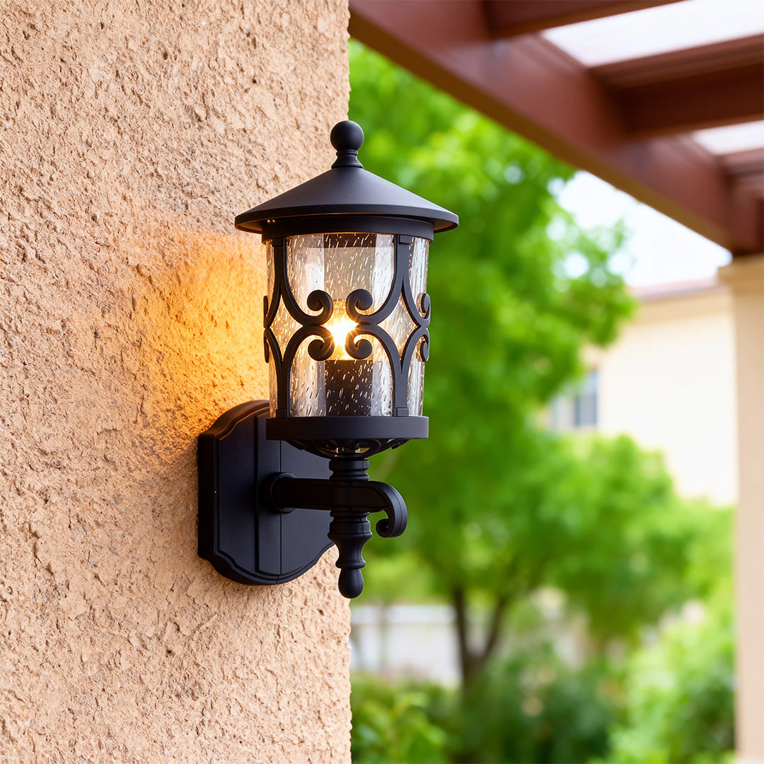 Outdoor black sconce casting warm glow on textured beige stucco wall with green foliage accent