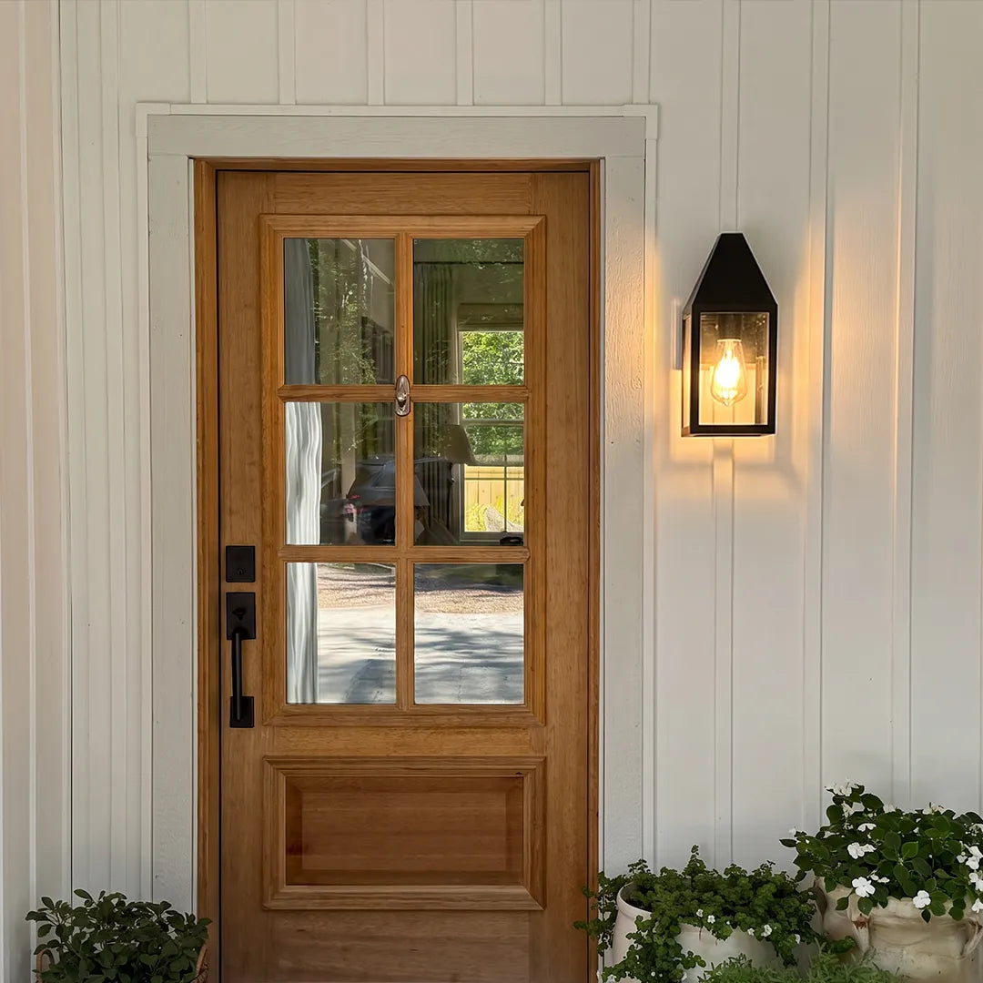 Outdoor black sconce installed beside a wooden front door on white siding, bringing classic exterior accent lighting.