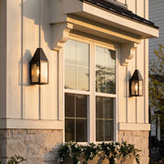 Outdoor black sconce mounted beside a window on a stone and stucco exterior, adding warm lantern-style lighting.