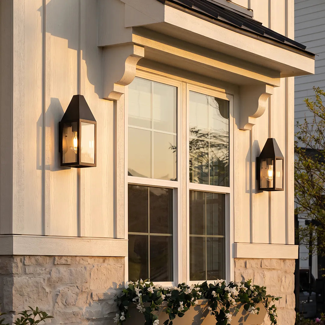 Outdoor black sconce mounted beside a window on a stone and stucco exterior, adding warm lantern-style lighting.