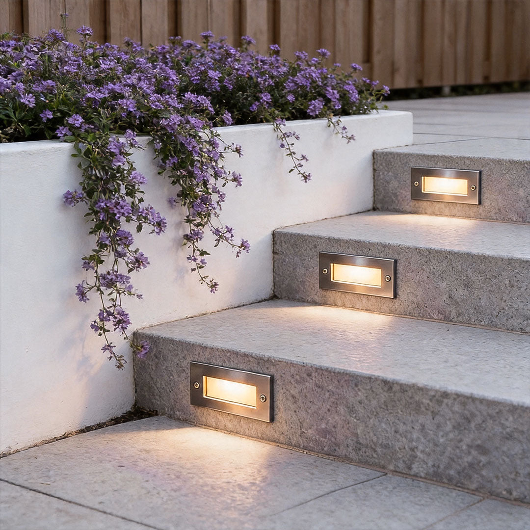 Outdoor brick light recessed into stone stair risers beside cascading purple flowers for elegant step illumination.