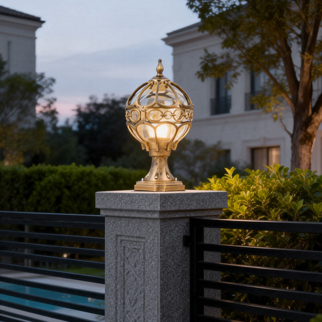 Outdoor globe pillar lights with intricate gold metalwork, mounted on a stone pillar near a modern house entrance.