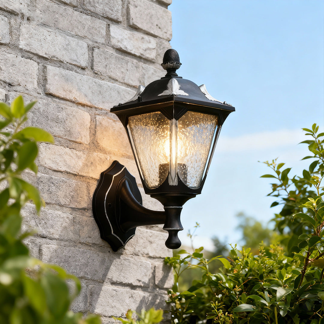 Outdoor light for wall casting warm glow on grey brick exterior near greenery and blue sky