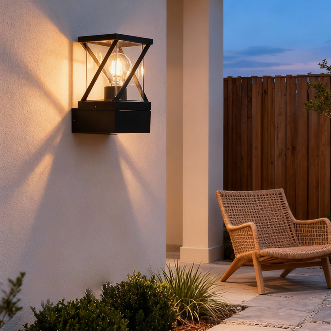 Warm-lit outdoor modern wall sconce on exterior wall near patio seating, featuring a cross-frame lantern structure.