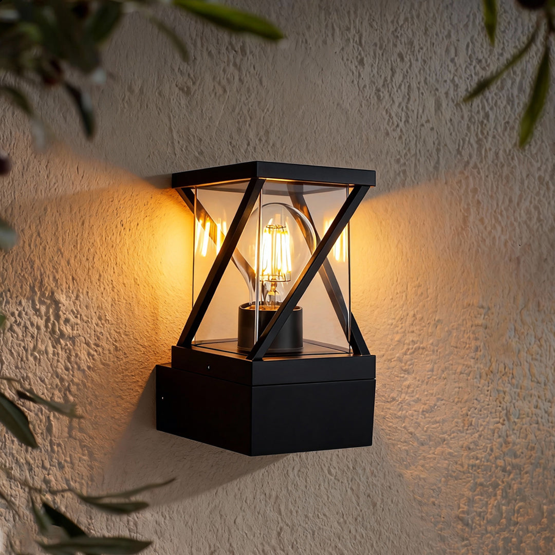 Warm glowing outdoor modern wall sconce mounted beside greenery, featuring a cross-frame lantern-style body.