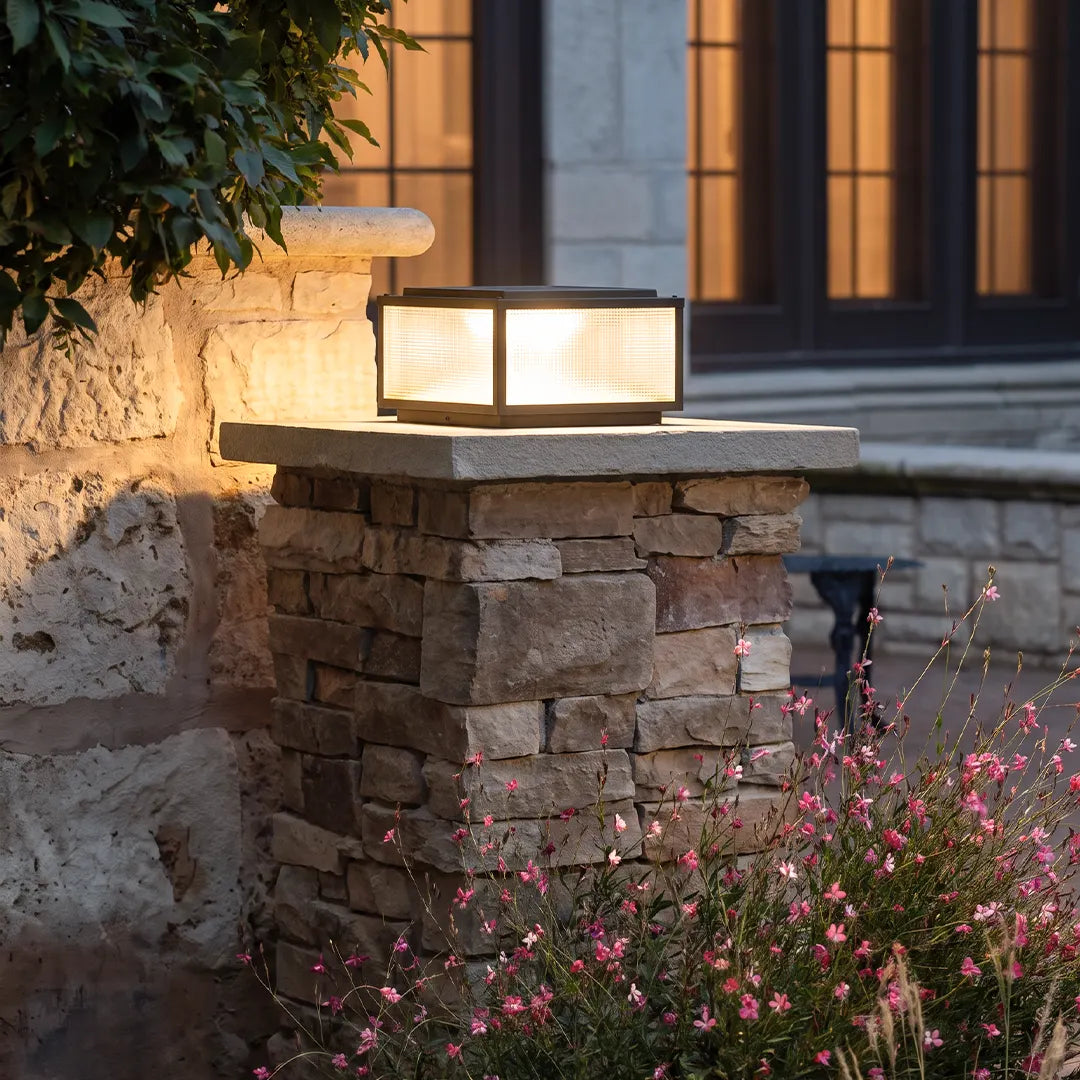 Outdoor pillar light fixture mounted on a stacked stone gate post beside a landscaped entry at dusk.