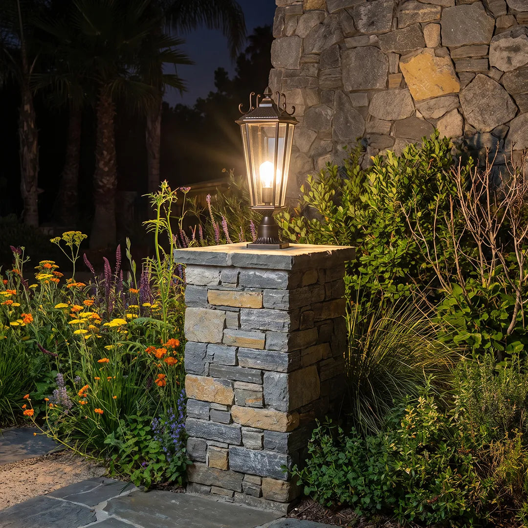 Outdoor pillar light fixtures on a stone landscape pillar casting a warm glow across shrubs and flowers at night.