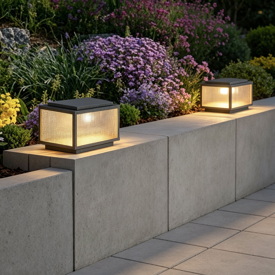 Outdoor pillar light fixtures arranged along a concrete landscape wall with flowering plants and pathway lighting.