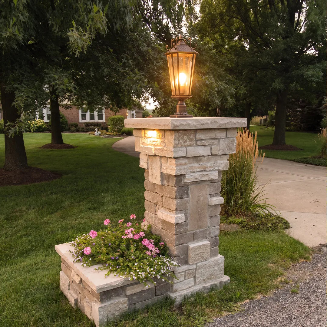 Outdoor pillar light fixtures installed on a stacked stone entry column with blooming flowers and warm evening illumination.
