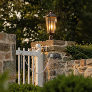 Outdoor pillar light fixtures mounted on a stone gate post beside a white garden fence in a landscaped yard.