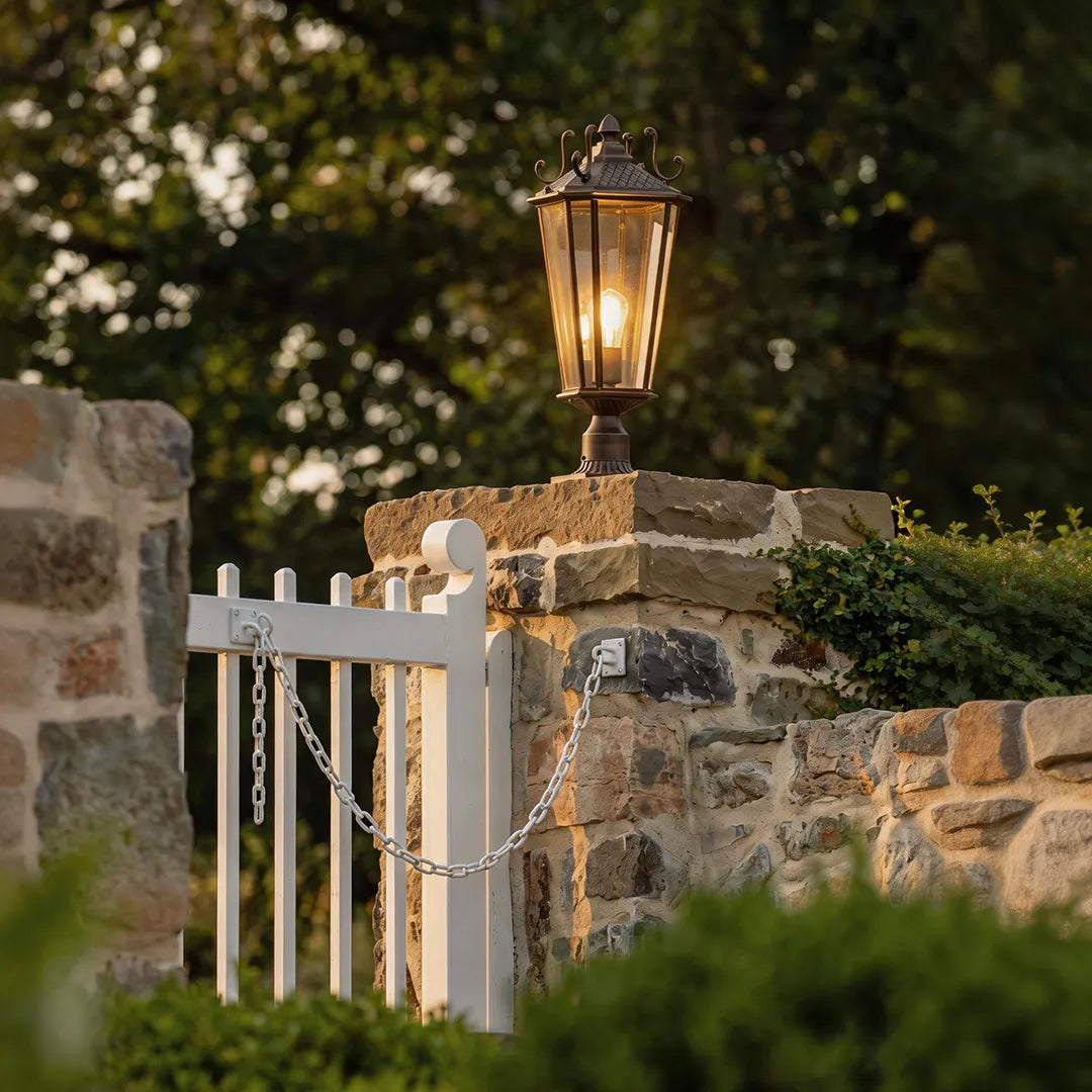Outdoor pillar light fixtures mounted on a stone gate post beside a white garden fence in a landscaped yard.