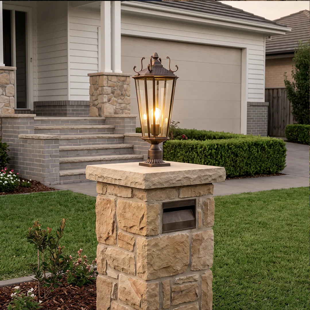 Outdoor pillar light fixtures displayed on a stone mailbox post in front of a suburban garage and lawn.