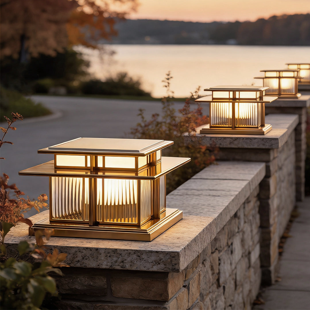 Outdoor porch pillar lights installed along a stone fence near a lakeside pathway at sunset.