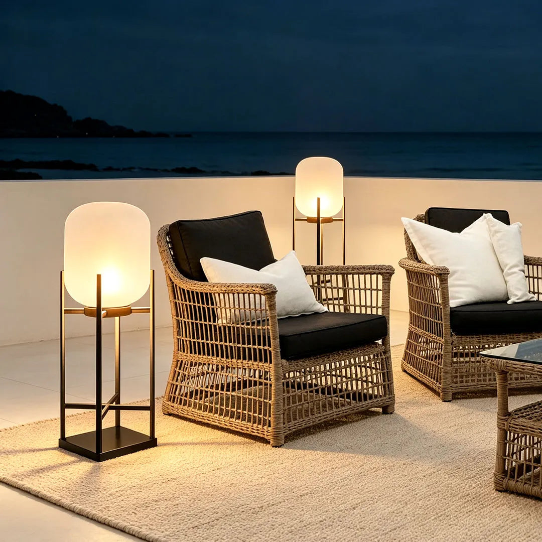 Outdoor standing light glowing beside wicker lounge seating on a coastal patio at night.