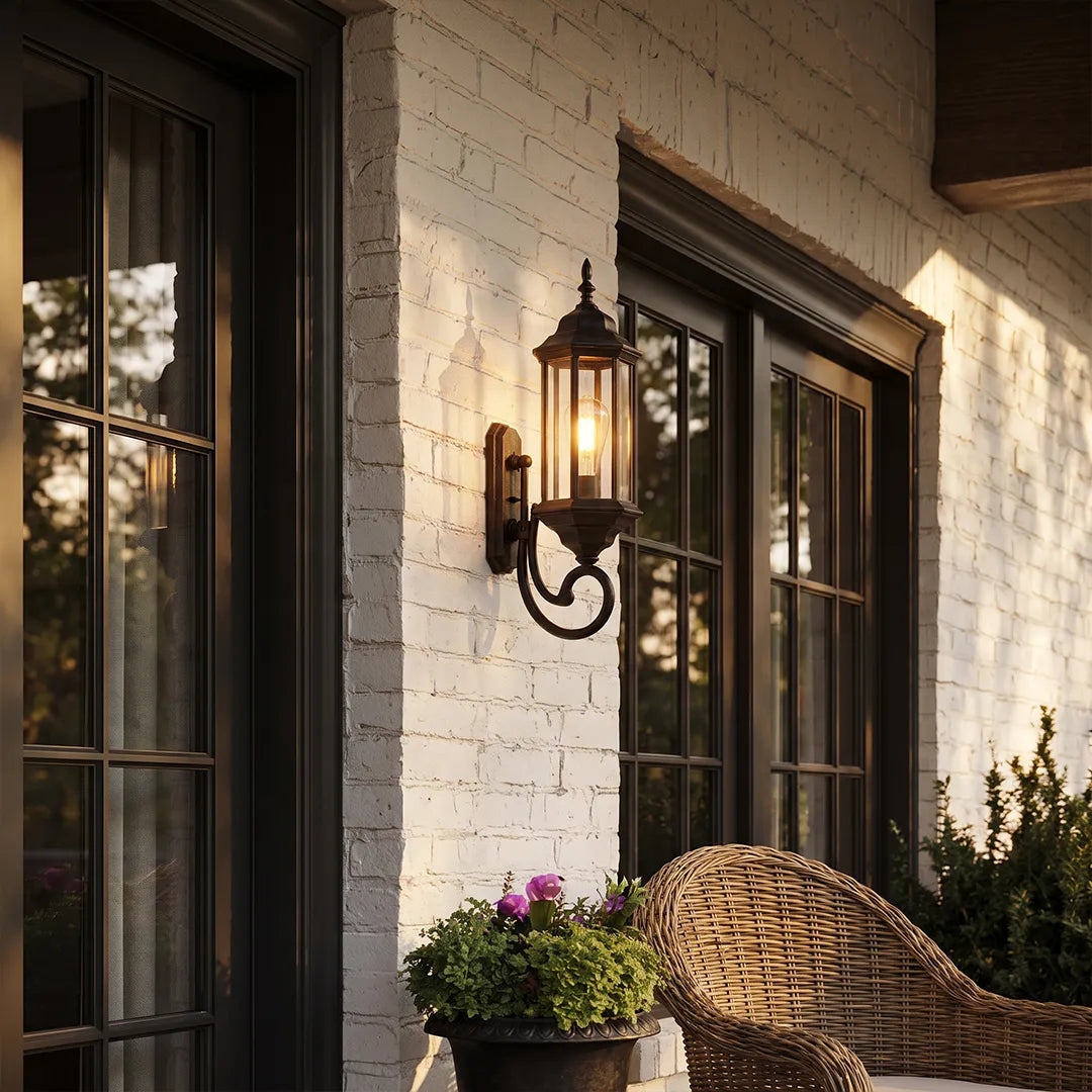 Outdoor wall lantern light on a brick exterior wall beside black framed windows and a woven patio chair.