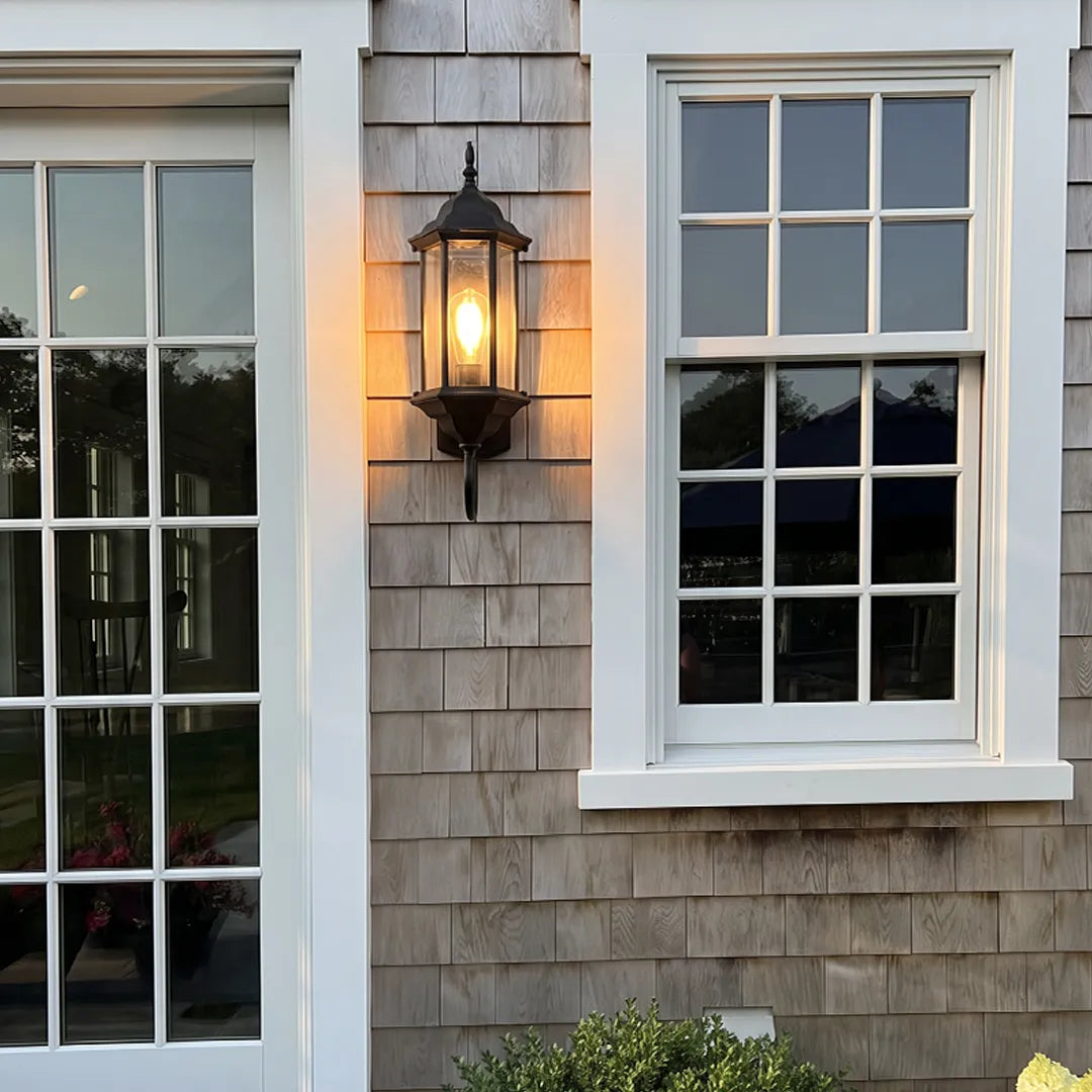 Outdoor wall lantern light mounted on cedar shake siding between white trimmed windows on a home exterior.