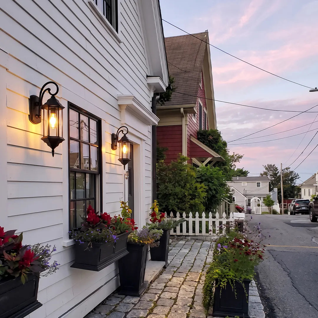 Outdoor wall lantern light installed on white siding along a side walkway with flower planters at dusk.