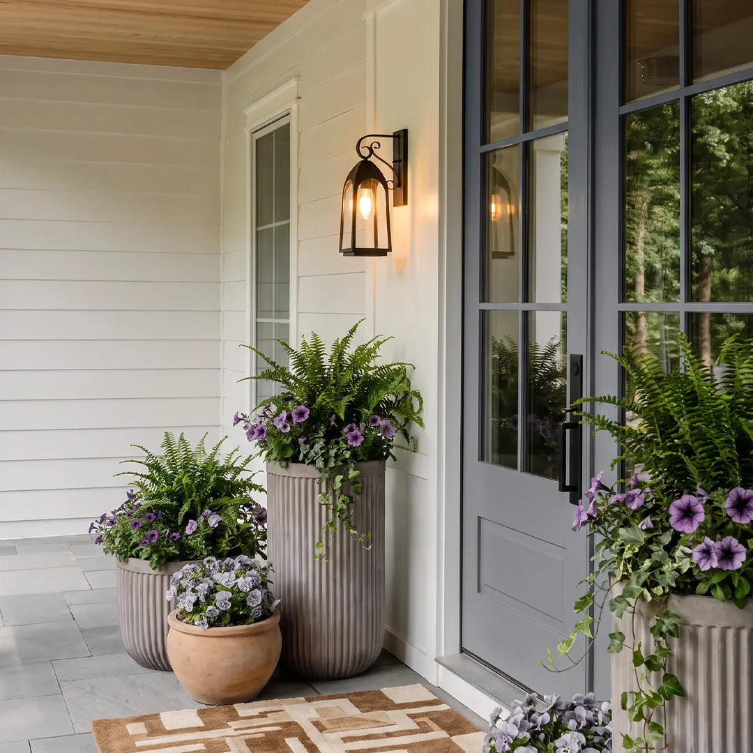 Outdoor wall mount lights by a front door with large planters and blooming purple flowers on a covered porch.