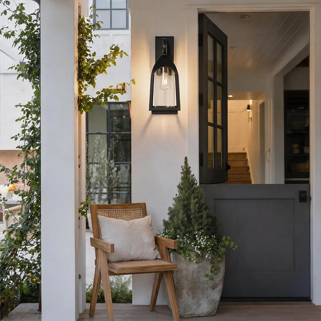 Outdoor wall mount lights beside a dark front door with a chair and potted plant on the porch.