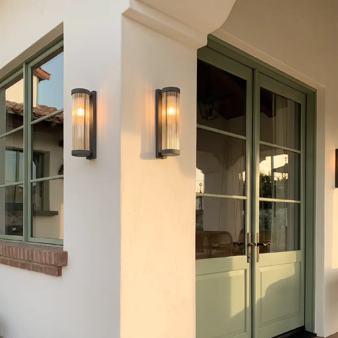 Outdoor wall mount lights mounted on a white exterior column beside green framed windows and doors, adding warm accent lighting.