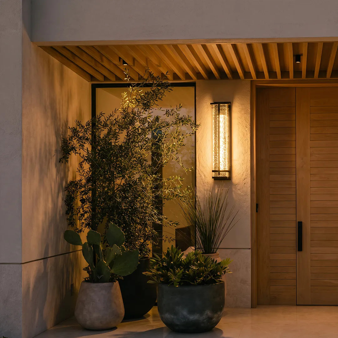 Outdoor wall sconce illuminating covered front porch with wood door and plants