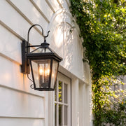Outdoor wall sconce light mounted on white exterior siding beside a window, featuring a black lantern frame and warm candle-style bulbs.