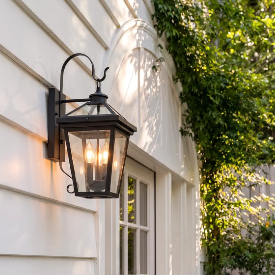 Outdoor wall sconce light mounted on white exterior siding beside a window, featuring a black lantern frame and warm candle-style bulbs.