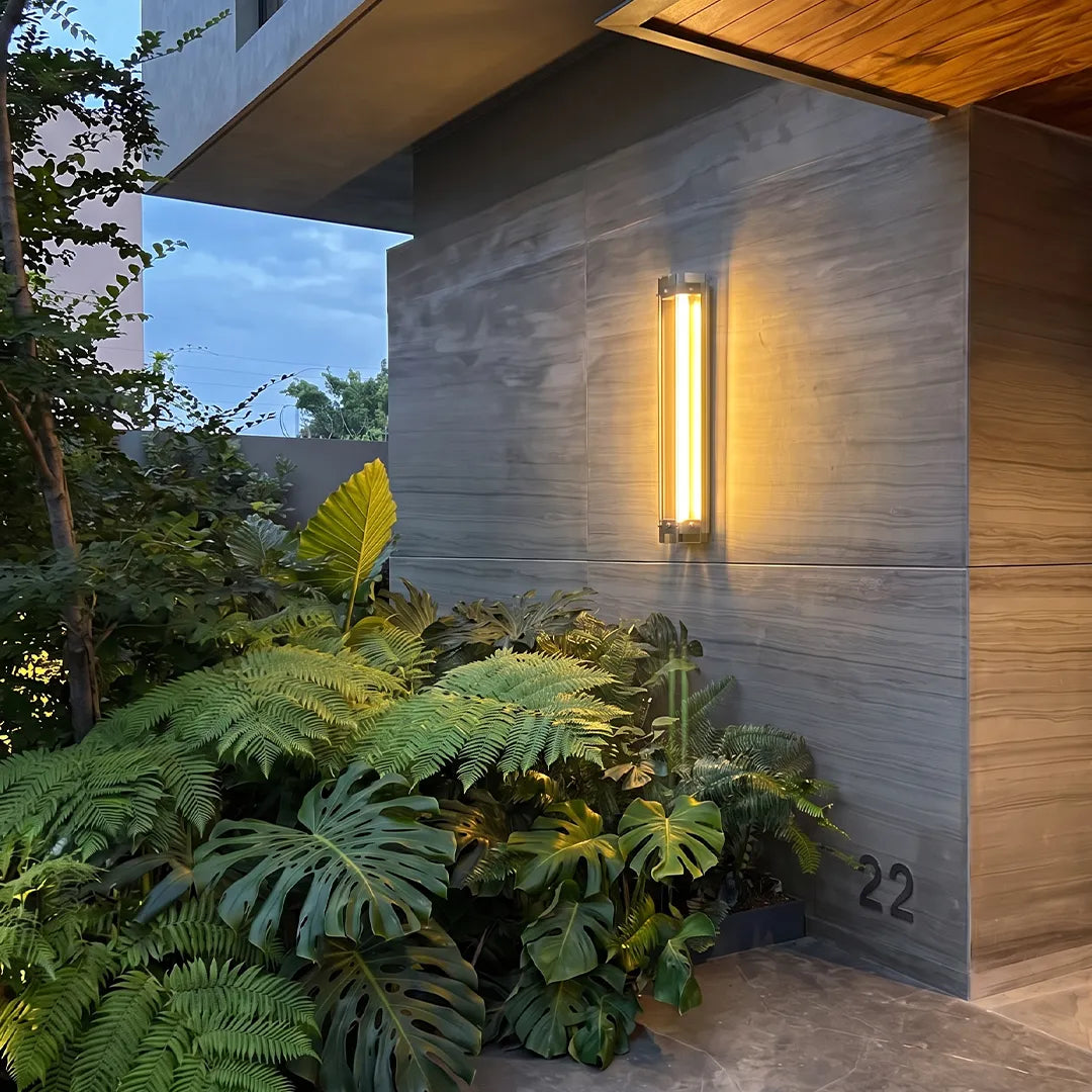 Outdoor wall sconce with warm glow on stone wall beside tropical garden