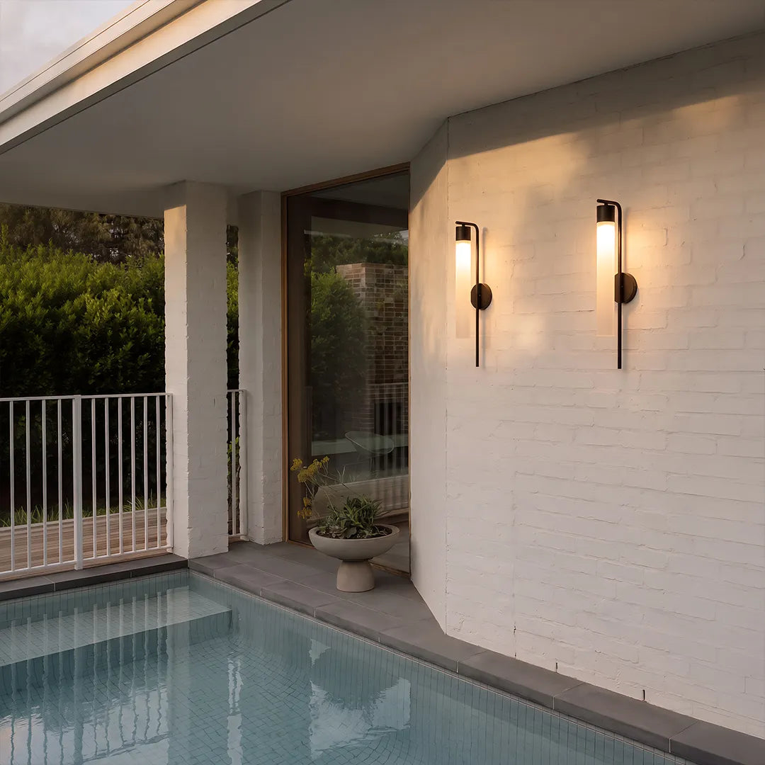 Outdoor wall sconces with warm glow on white brick wall beside swimming pool