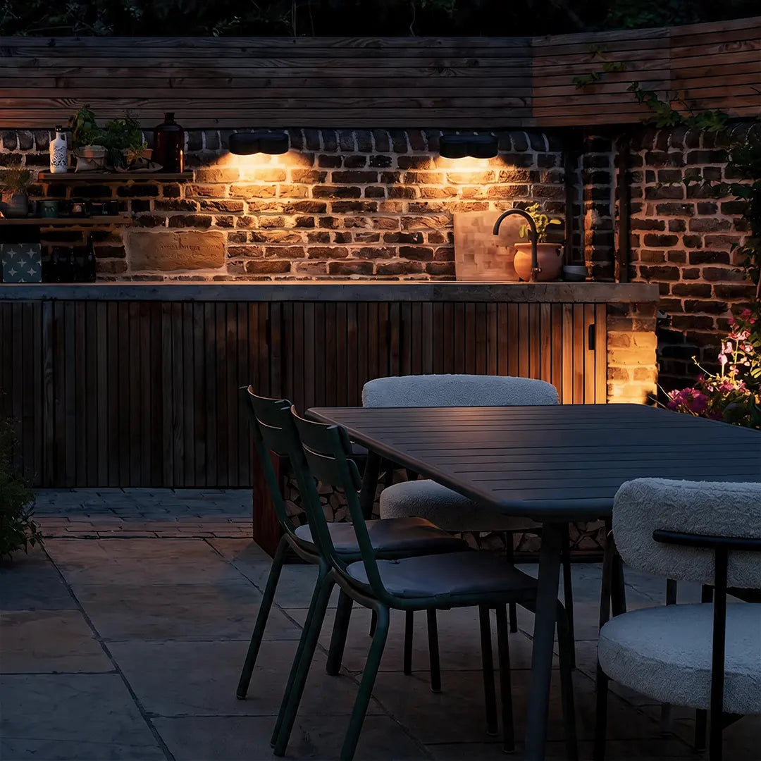 Outdoor wall wash lighting illuminating a brick patio dining area and outdoor kitchen wall at night.