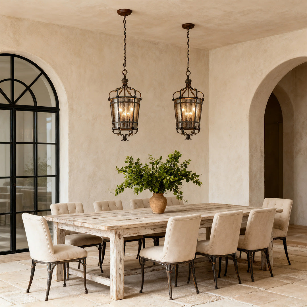 Pair of Italian pendant light fixtures with aged bronze caged design, suspended over a rustic, light wood dining table in a Mediterranean-style room.
