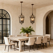Pair of Italian pendant light fixtures with aged bronze caged design, suspended over a rustic, light wood dining table in a Mediterranean-style room.