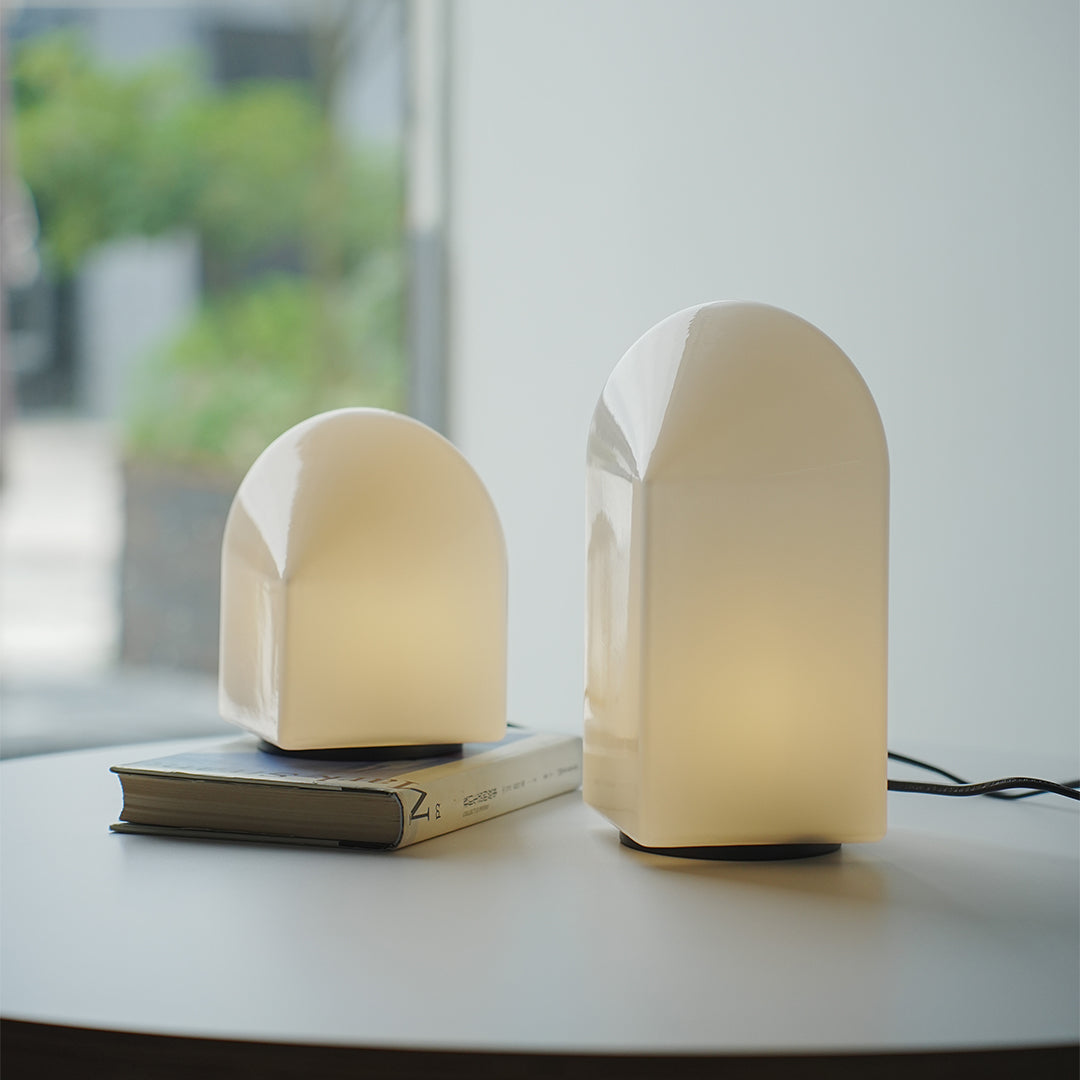 Pair of house-shaped modern white table lamps in different sizes on round black table.