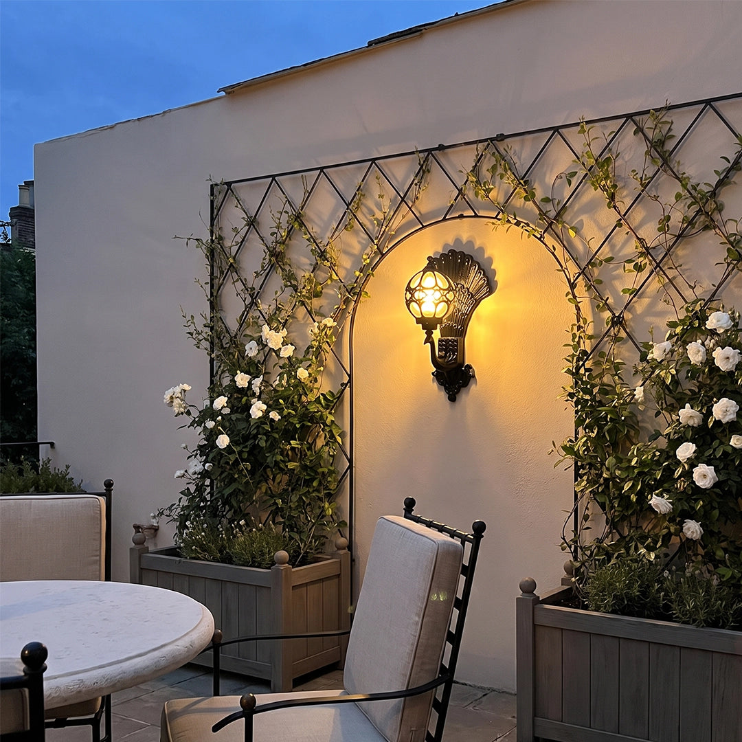 Peacock wall light displayed in an arched garden wall niche, casting a warm glow over an outdoor patio setting.