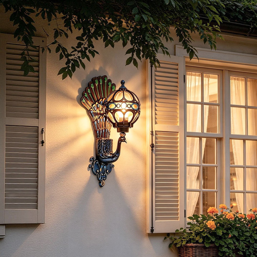 Peacock wall light with an ornate fan-shaped backplate and glowing globe shade, mounted beside a shuttered window on an exterior wall.
